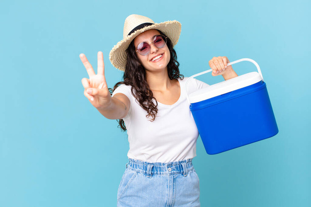 pretty hispanic woman smiling and looking friendly, showing number two holding a portable refrigerator - Foto, Imagen