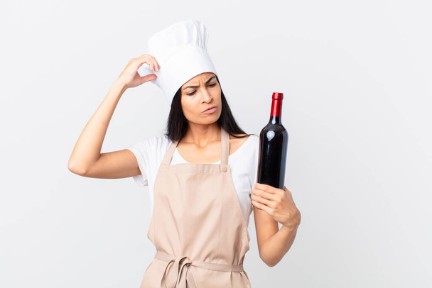 pretty hispanic chef woman feeling puzzled and confused, scratching head and holding a bottle of wine - Fotó, kép