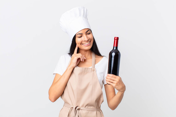 pretty hispanic chef woman smiling happily and daydreaming or doubting and holding a bottle of wine - Fotoğraf, Görsel