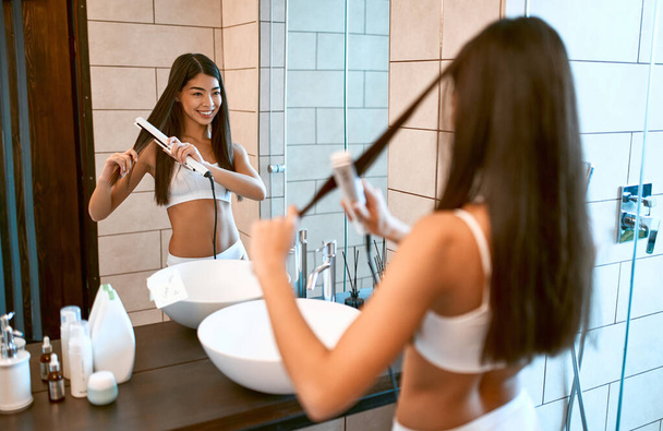 Egy koreai nő fehér alsóneműben a tükör előtt a fürdőszobában kiegyenesíti a vashajat, megcsinálja a stílust.. - Fotó, kép