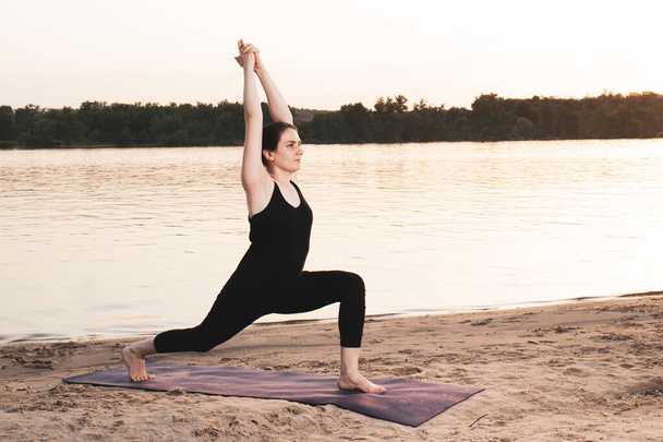 Una mujer practica yoga mientras está de pie en la posición de Guerrero 1 o Virabhadrasana Uno. Yoga al atardecer en la naturaleza cerca del río. - Foto, imagen