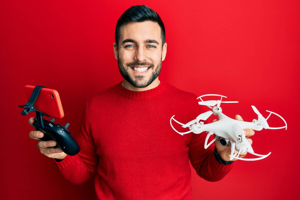 Νεαρός Ισπανόφωνος κρατώντας drone και τηλεχειριστήριο με smartphone χαμογελώντας με ένα χαρούμενο και δροσερό χαμόγελο στο πρόσωπο. που δείχνει δόντια.  - Φωτογραφία, εικόνα