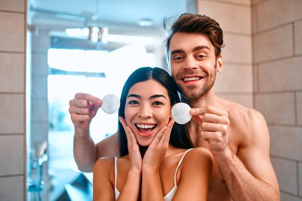 A couple in love in the bathroom have fun. A man funnyly holds cotton discs to his girlfriend. Removing make-up. Facial skin care. - Photo, Image