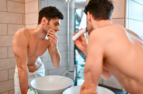 A beautiful muscular man in a white towel stands in the bathroom in front of a mirror and shaves his face. Caring for the face and body. - Фото, зображення