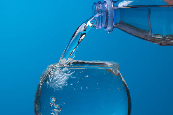 Νερό χύνεται σε ένα ποτήρι από ένα πλαστικό μπουκάλι σε μπλε φόντο. Καθαρό πόσιμο νερό. - Φωτογραφία, εικόνα