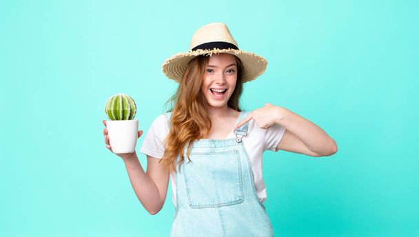cabeza roja bonita campesina sintiéndose feliz y señalándose a sí misma con una excitada y sosteniendo un cactus - Foto, Imagen