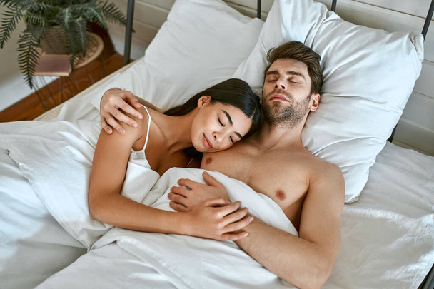 Утро влюбленной молодой пары. Красивая корейская женщина и мускулистый мужчина лежат под одеялом в кровати в светлой современной спальне. - Фото, изображение