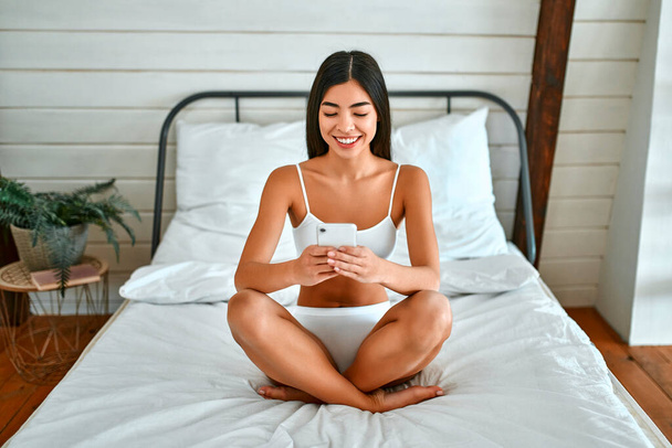 Een prachtige Koreaanse vrouw in wit ondergoed in de slaapkamer zit op het bed en schrijft een bericht op haar smartphone. Ochtend een prachtig slank meisje in een witte slaapkamer. - Foto, afbeelding