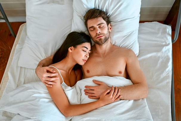 Утро влюбленной молодой пары. Красивая корейская женщина и мускулистый мужчина лежат под одеялом в кровати в светлой современной спальне. - Фото, изображение