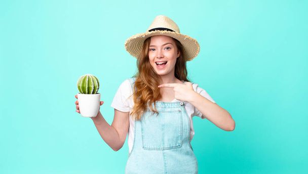 red head pretty farmer woman looking excited and surprised pointing to the side and holding a cactus - Фото, изображение