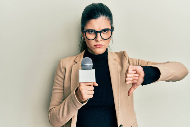 Молодая латиноамериканка держит в руках репортера микрофон с сердитым лицом, негативный знак, показывающий неприязнь с опущенными пальцами, концепция отказа  - Фото, изображение