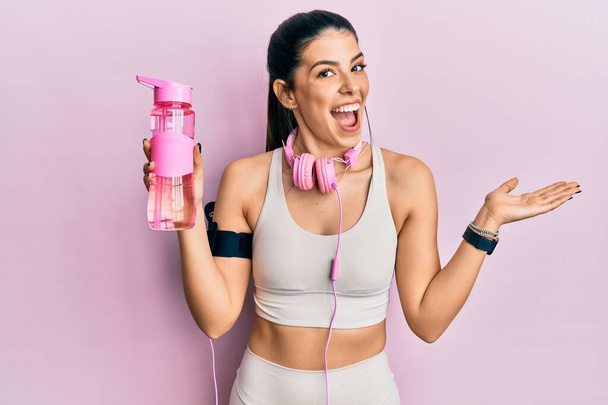 Junge hispanische Frau in Sportkleidung, Trinkflasche mit Wasser und Kopfhörer, die Leistung mit glücklichem Lächeln und Siegerausdruck mit erhobener Hand feiert  - Foto, Bild