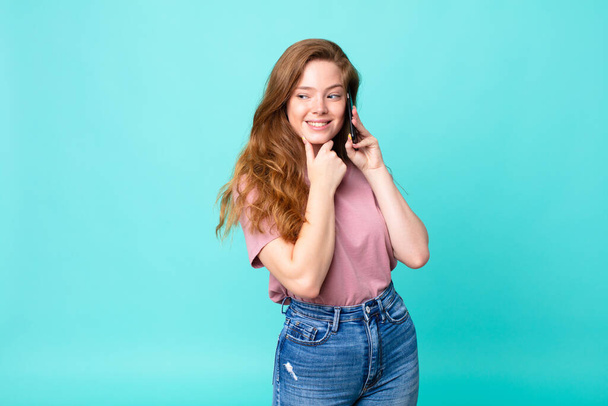 red head pretty woman smiling with a happy, confident expression with hand on chin and using a smartphone - Foto, Imagen