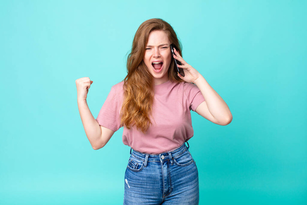red head pretty woman shouting aggressively with an angry expression and using a smartphone - Fotoğraf, Görsel