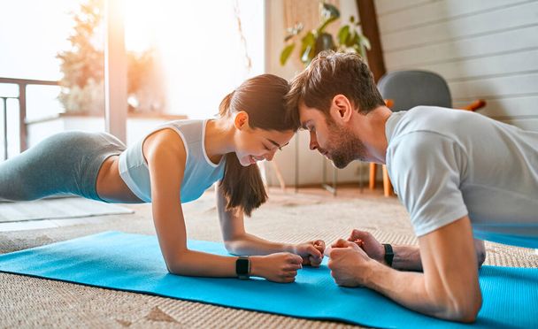 Een leuk stel in sportkleding doet een oefenplankje op de matten in de woonkamer thuis. Gezonde levensstijl, sport, yoga, fitness. - Foto, afbeelding