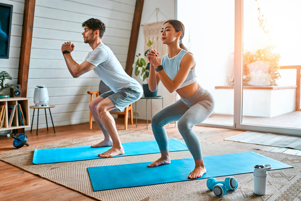 Cute couple woman Korean and muscular man in sports clothes do exercises squat on mats in the living room at home. Healthy lifestyle, yoga, sports, fitness. - Photo, Image