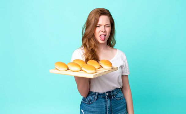 red head pretty woman feeling disgusted and irritated and tongue out and holding a bread buns troy - Foto, Imagem
