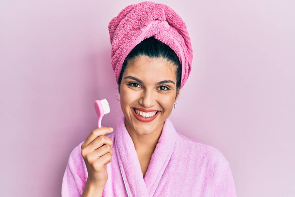 Mulher hispânica jovem vestindo roupão de banho segurando escova de dentes olhando positivo e feliz de pé e sorrindo com um sorriso confiante mostrando dentes  - Foto, Imagem