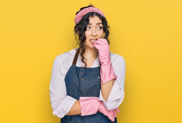 Молода іспаномовна дівчина носить чистіший фартух і рукавички, які виглядають напруженими і нервують руками на роті, кусаючи цвяхи. проблема тривоги
.  - Фото, зображення
