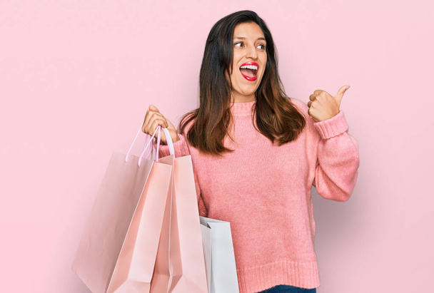 Hermosa mujer hispana sosteniendo bolsas de compras apuntando con el pulgar hacia el lado sonriendo feliz con la boca abierta  - Foto, imagen