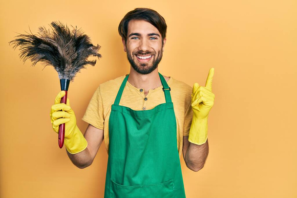 Молодий іспаномовний чоловік у фартуху тримає прибиральник, усміхаючись щасливим вказуючи рукою і пальцем на бік
  - Фото, зображення
