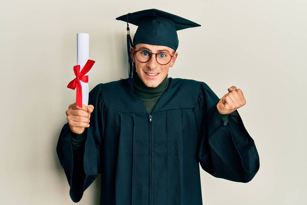 Young caucasian man wearing graduation cap and ceremony robe holding diploma screaming proud, celebrating victory and success very excited with raised arm  - Φωτογραφία, εικόνα