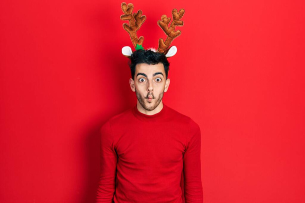 Young hispanic man wearing cute christmas reindeer horns making fish face with lips, crazy and comical gesture. funny expression.  - Photo, Image