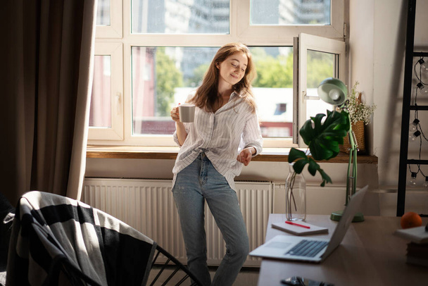 Young beautiful brunette woman drinking morning coffee while standing by the window in the apartment near table with laptop - Фото, зображення