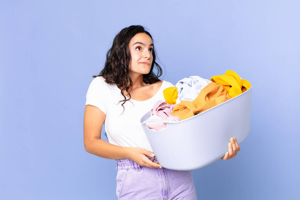 hispanic pretty woman shrugging, feeling confused and uncertain and holding a washing clothes basket - Foto, immagini