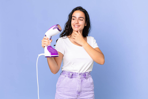 hispanic pretty woman smiling with a happy, confident expression with hand on chin and holding an iron steam - Zdjęcie, obraz