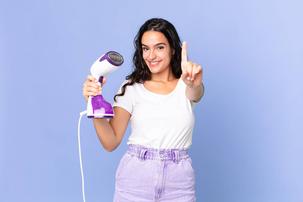 hispanic pretty woman smiling proudly and confidently making number one and holding an iron steam - Photo, Image