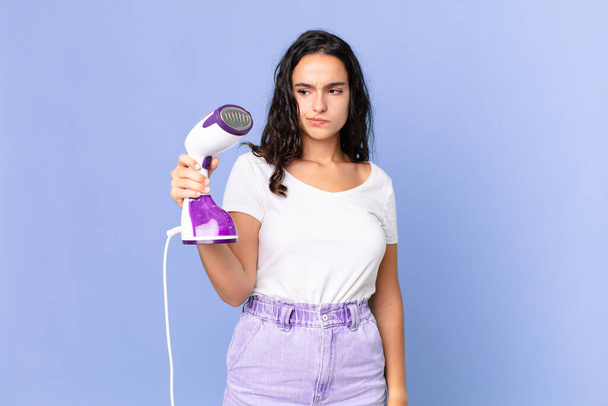 hispanic pretty woman feeling sad, upset or angry and looking to the side and holding an iron steam - Photo, image