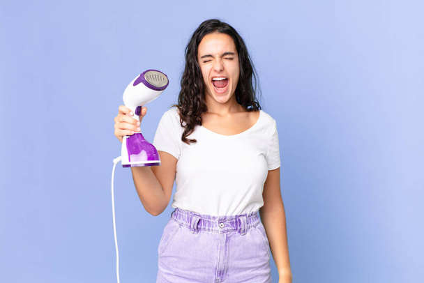 hispanic pretty woman shouting aggressively, looking very angry and holding an iron steam - Photo, image