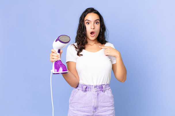hispanic pretty woman looking shocked and surprised with mouth wide open, pointing to self and holding an iron steam - Photo, Image