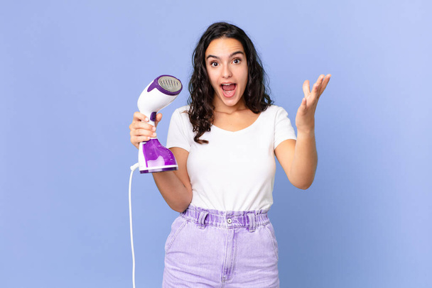 hispanic pretty woman feeling extremely shocked and surprised and holding an iron steam - Фото, зображення
