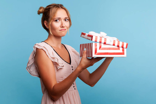 Portrait of upset young adult blonde female with bundles, holding red striped present box, has sad expression, being disappointed with birthday gift. Indoor studio shot isolated on blue background. - Foto, immagini