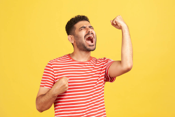 Extremely excited overjoyed man with beard in striped t-shirt shouting making yes gesture, amazed with his victory, triumph. Indoor studio shot isolated on yellow background - Foto, Bild