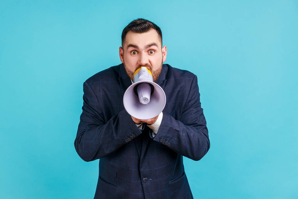 Portrait of angry nervous bearded man wearing elegant suit loudly screaming at megaphone, making announce, protesting, wants to be heard. Indoor studio shot isolated on blue background. - Фото, изображение