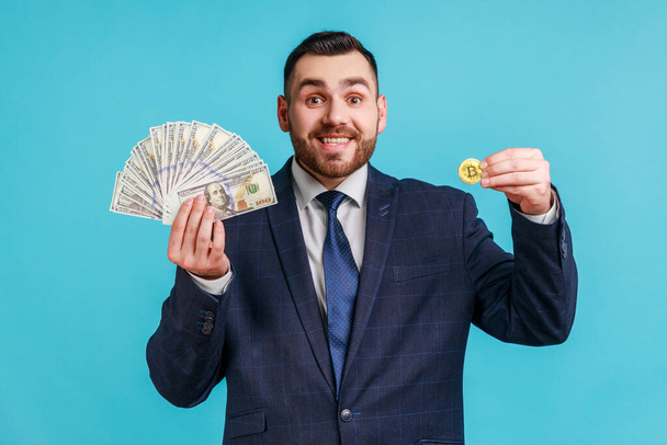 Cryptocurrency btc and money. Portrait of rich happy bearded man elegant suit showing dollar banknotes and golden bitcoin, smiling to camera. Indoor studio shot isolated on blue background. - Фото, изображение