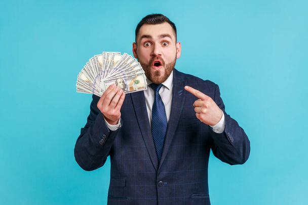 Wow look, I am rich! Surprised bearded man in suit showing and pointing at dollar banknotes, staring at camera in amazement, shocked by business profit. Indoor studio shot isolated on blue background. - Фото, изображение