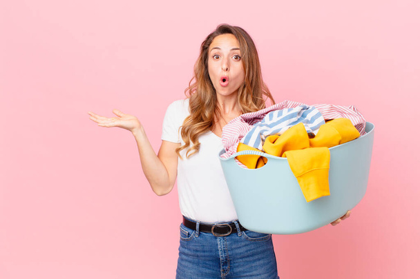pretty woman looking surprised and shocked, with jaw dropped holding an object and washing clothes. - Foto, Bild