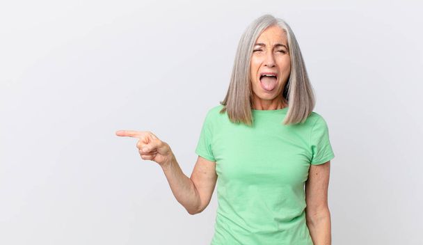 middle age white hair woman with cheerful and rebellious attitude, joking and sticking tongue out and pointing to the side - Foto, Bild