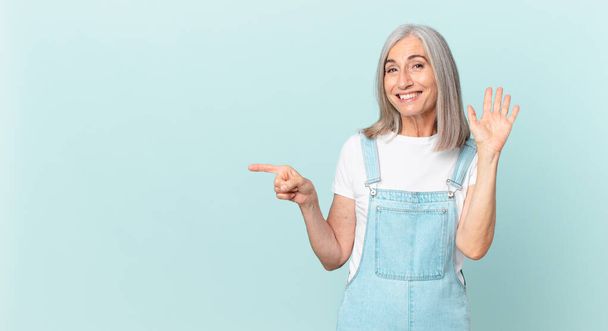 middle age white hair woman smiling happily, waving hand, welcoming and greeting you and pointing to the side - Foto, Imagen