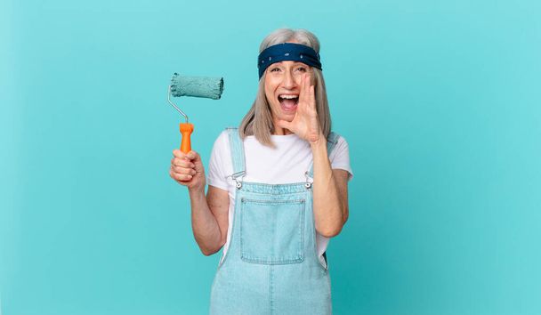 middle age white hair woman feeling happy,giving a big shout out with hands next to mouth with a roller painting a wall - Fotoğraf, Görsel
