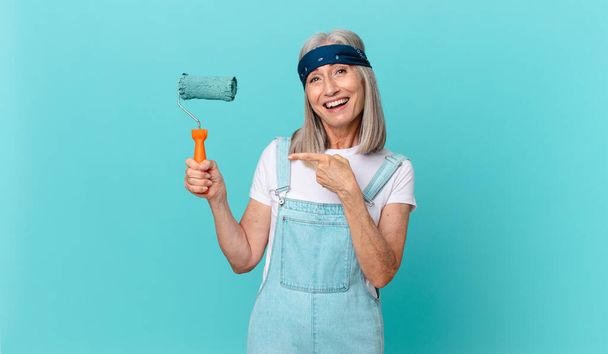 middle age white hair woman looking excited and surprised pointing to the side with a roller painting a wall - Photo, image