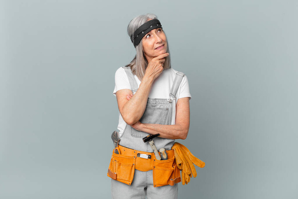 middle age white hair woman smiling with a happy, confident expression with hand on chin and wearing work wear and tools. housekeeping concept - Fotó, kép