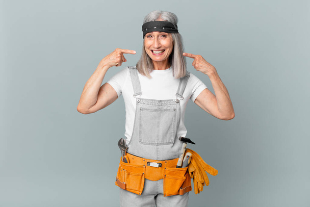 middle age white hair woman smiling confidently pointing to own broad smile and wearing work wear and tools. housekeeping concept - Fotografie, Obrázek
