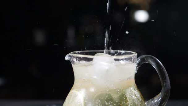 Šumivá limonádová voda se nalévá do skleněné nádoby naplněné plátky vápna, mátových lístků a ledu, stříkance. Letní ovoce a bobule. Osvěžující koktejl na černém pozadí s kostkami ledu na stole. Pomalé - Záběry, video