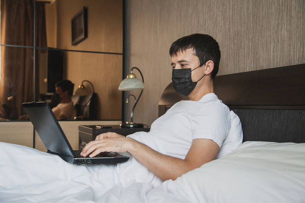 Mies istuu sängyssä ja työskentelee kannettavalla tietokoneella, jolla on lääketieteellinen naamio kasvoillaan.. - Valokuva, kuva