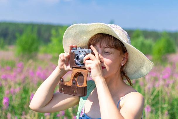 Portrait of a young woman with a camera in a hat in a field of red flowers on a hot summer day. Selective focus. Close-up - Фото, изображение